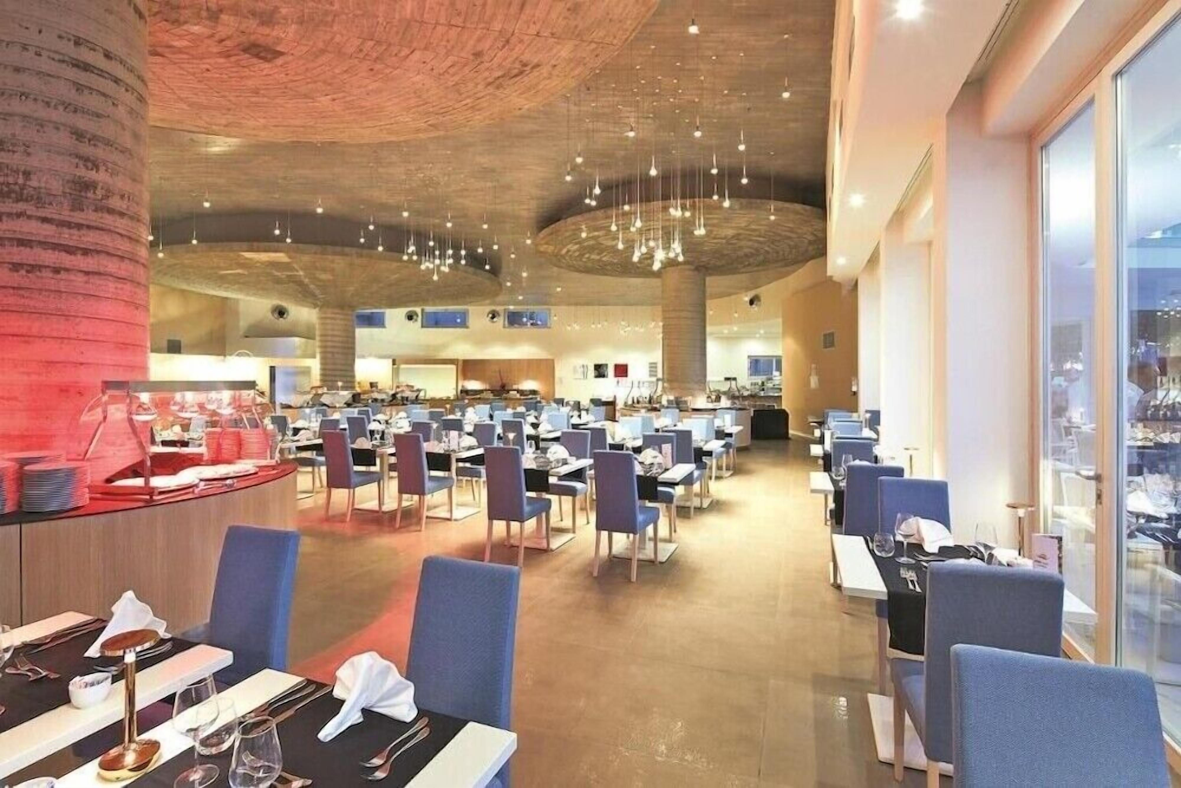 A spacious restaurant with multiple dining tables and chairs, a high ceiling with hanging lights, and a view through large windows.