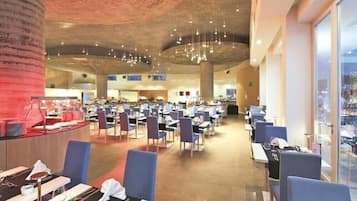 A spacious restaurant with multiple dining tables and chairs, a high ceiling with hanging lights, and a view through large windows.