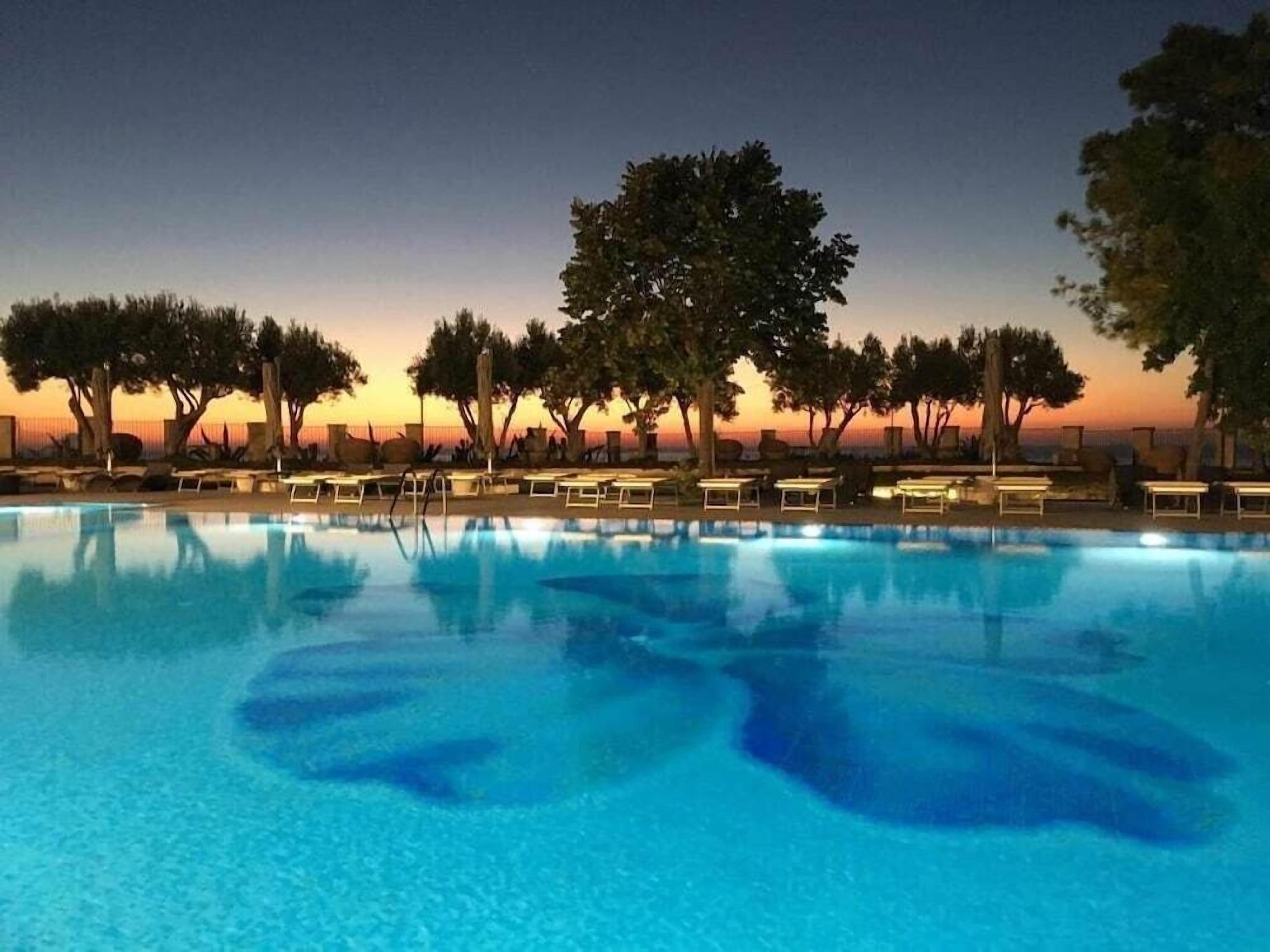 A swimming pool with lounge chairs and trees in the background during twilight.