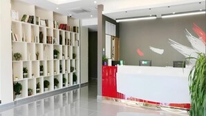 A modern reception area with a white reception desk, shelves with books and plants, and a grey wall with abstract art. - Thank (Shanghai)