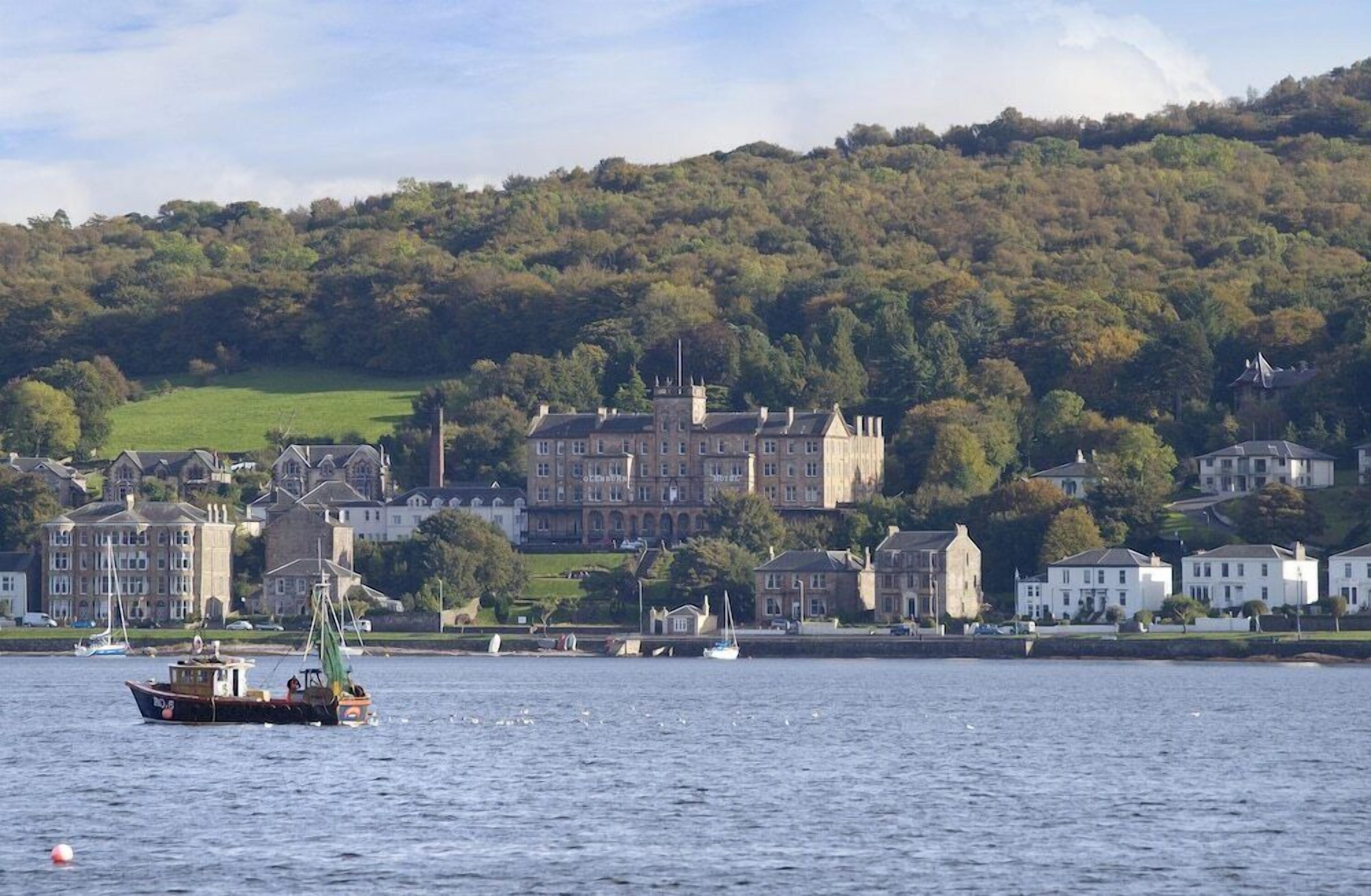A coastal town with a prominent castle-like building, a fishing boat in the foreground, and a hilly backdrop.