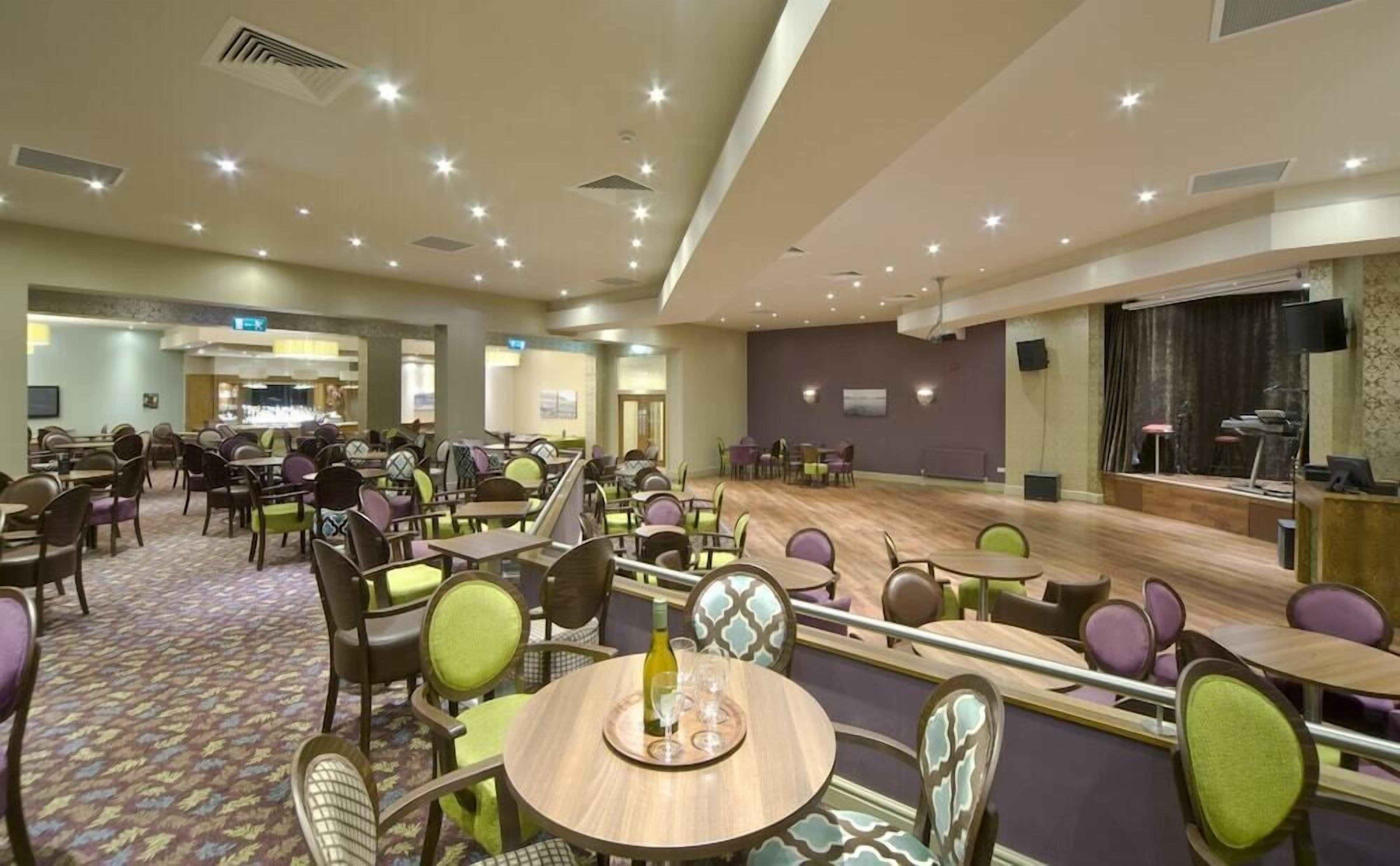 Un amplio salón comedor de hotel con varias mesas y sillas, un escenario y una zona de bar.