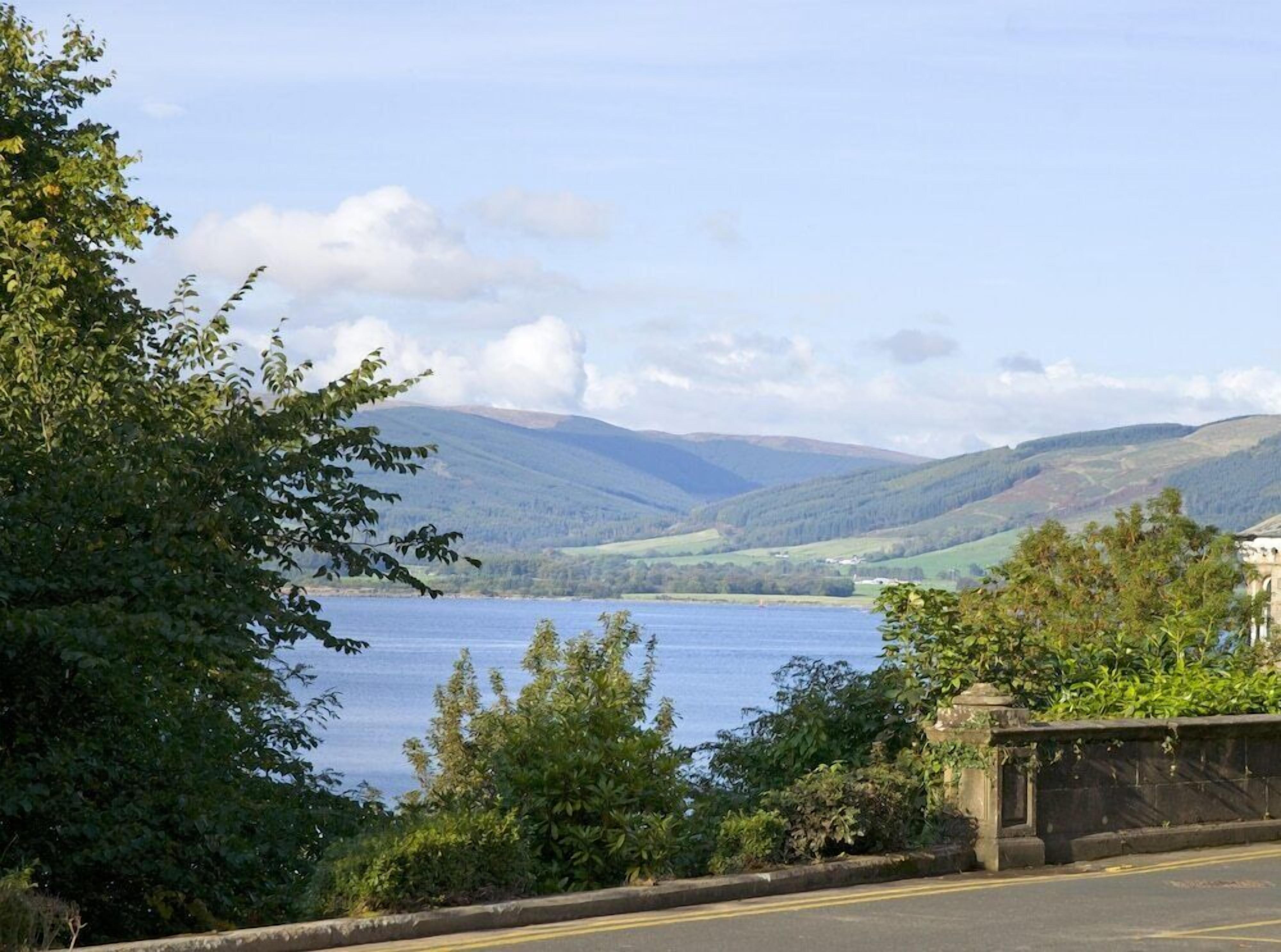A scenic view of a lake with hills in the background, a road with a stone wall on the right, and lush greenery on the left.