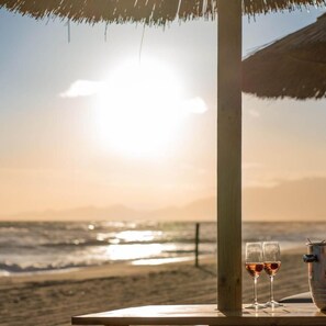 A beach scene with a table under a thatched roof, two glasses of drink, and a metal container. - Cumeja Beach Club Hotel (Cellole)