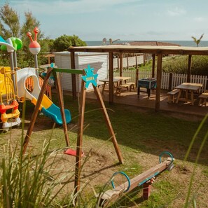 A playground with a slide and swings, a covered seating area with tables, and a view of the sea. - Cumeja Beach Club Hotel (Cellole)