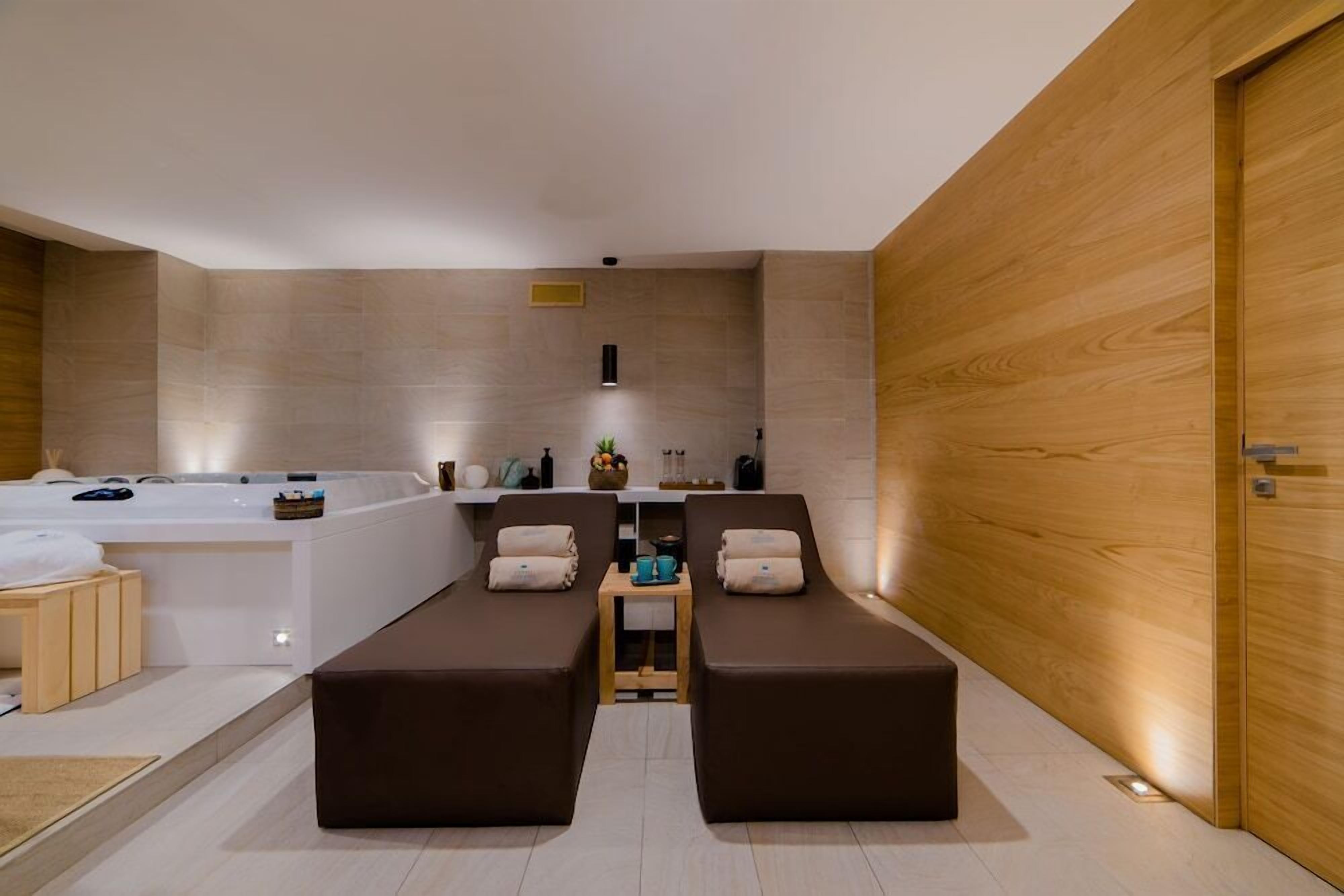 Un'elegante sala spa moderna con una grande vasca da bagno, due poltrone reclinabili, un tavolino con articoli da toilette e una parete in legno.