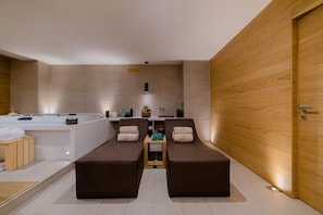 Een moderne spa-ruimte met een groot bad, twee relaxstoelen, een klein tafeltje met verzorgingsproducten en een houten wand.