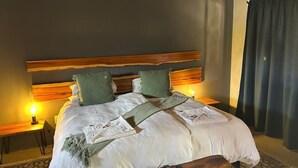 A neatly made bed with a wooden headboard, two pillows, and a blanket. There are bedside tables with lamps and a remote control on the bed. - Klein Karoo Game Lodge (De Hoop)
