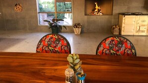 A dining area with a wooden table, two chairs with floral upholstery, a fireplace, and a window with a view of trees. - Klein Karoo Game Lodge (De Hoop)