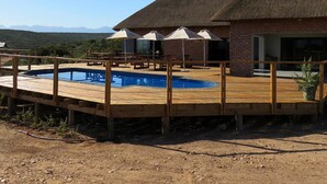 A pool with a wooden deck, a building with a thatched roof, and a clear sky. - Klein Karoo Game Lodge (De Hoop)