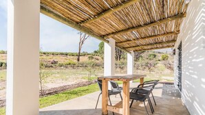 Eine überdachte Terrasse mit Betonboden, einem Holztisch und zwei schwarzen Stühlen.