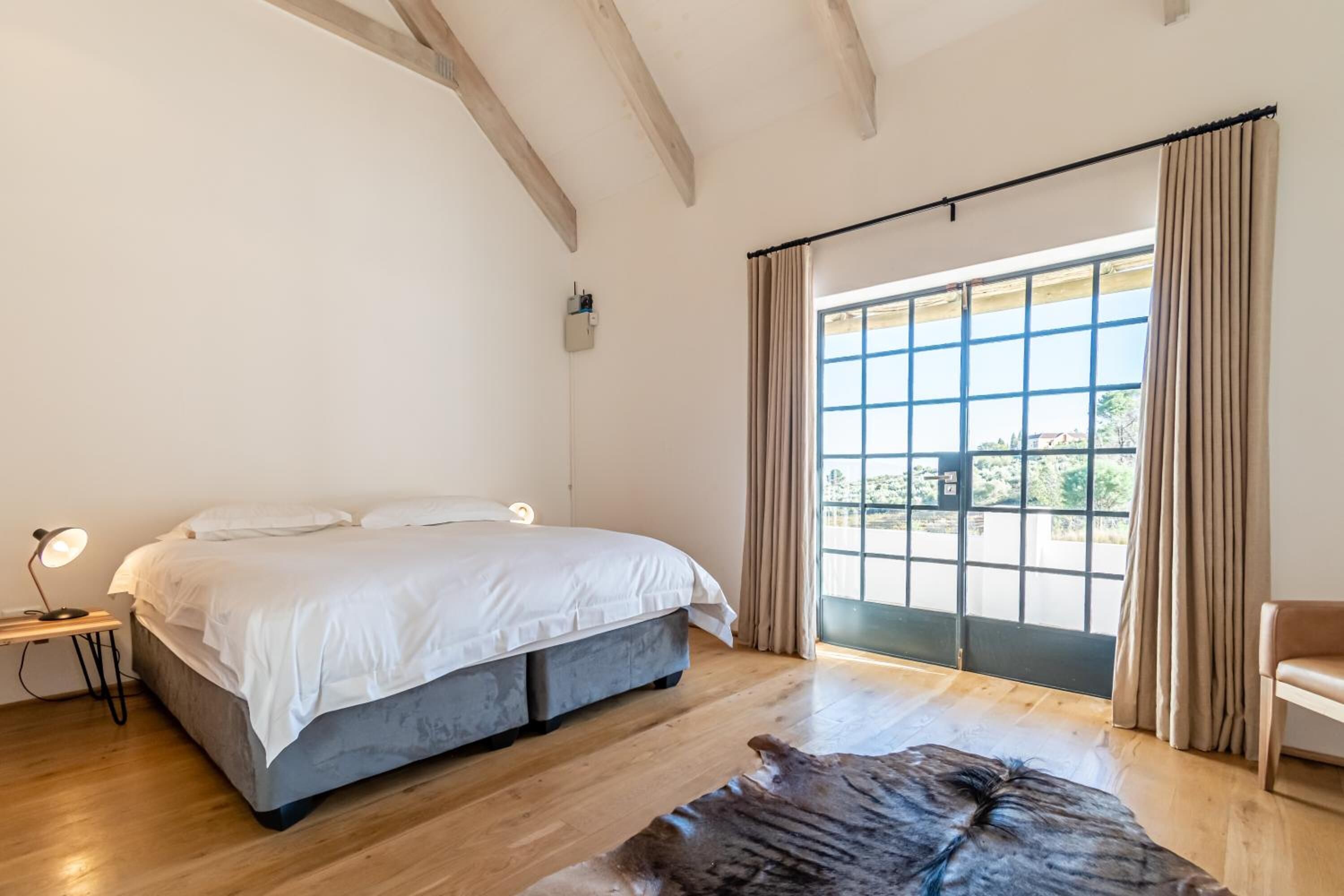 A bedroom with a bed, a desk with a lamp, a leather rug, and a large window with curtains.