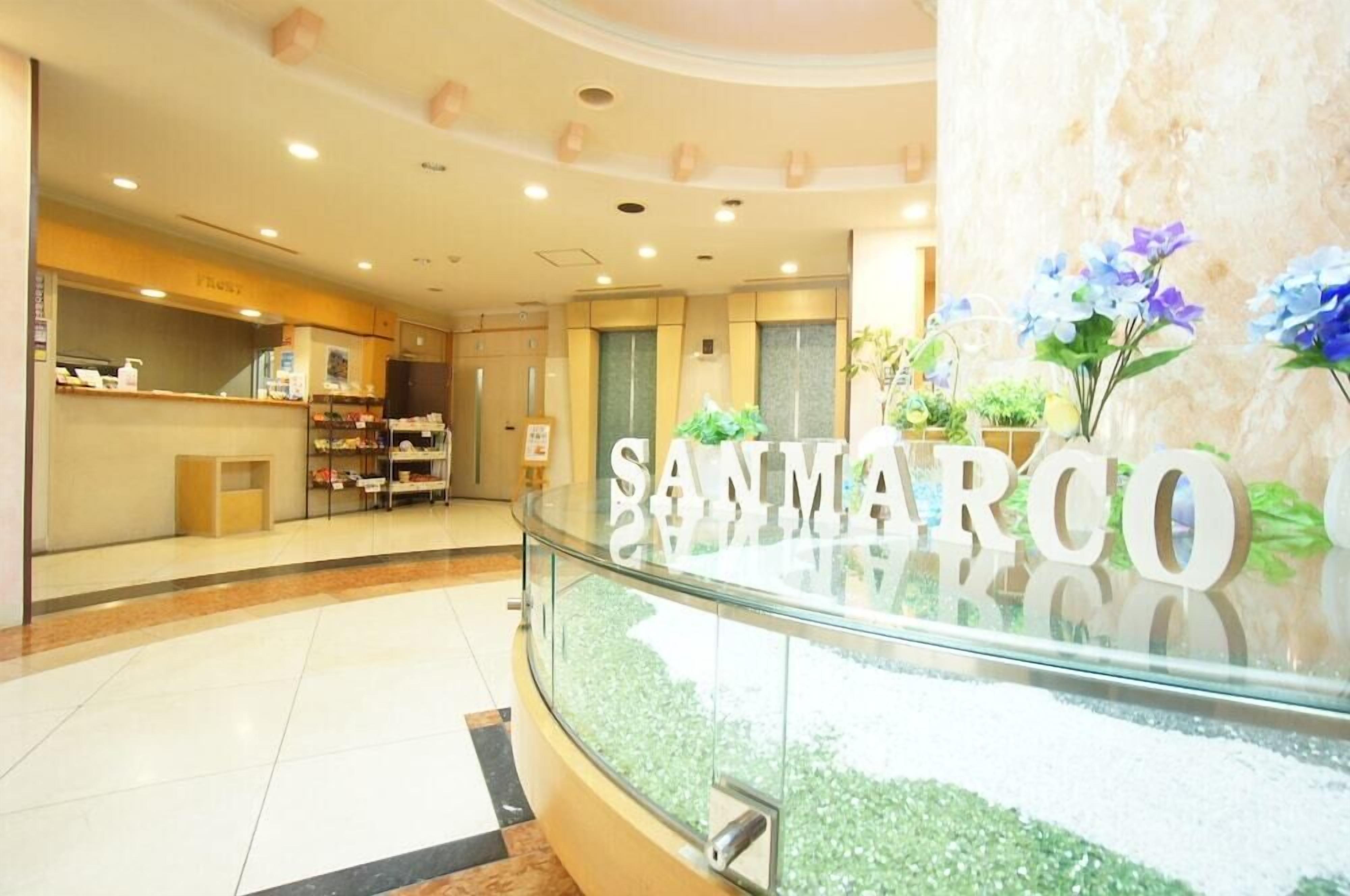 A hotel lobby with a reception desk, a glass railing, and a sign that reads "SANMARCO".