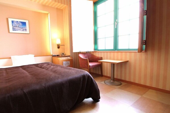 Ein Hotelzimmer mit Bett, einem kleinen Tisch, einem Stuhl, einem Nachttisch, einer Lampe, einem Fenster mit Vorhängen und einem gerahmten Bild an der Wand.