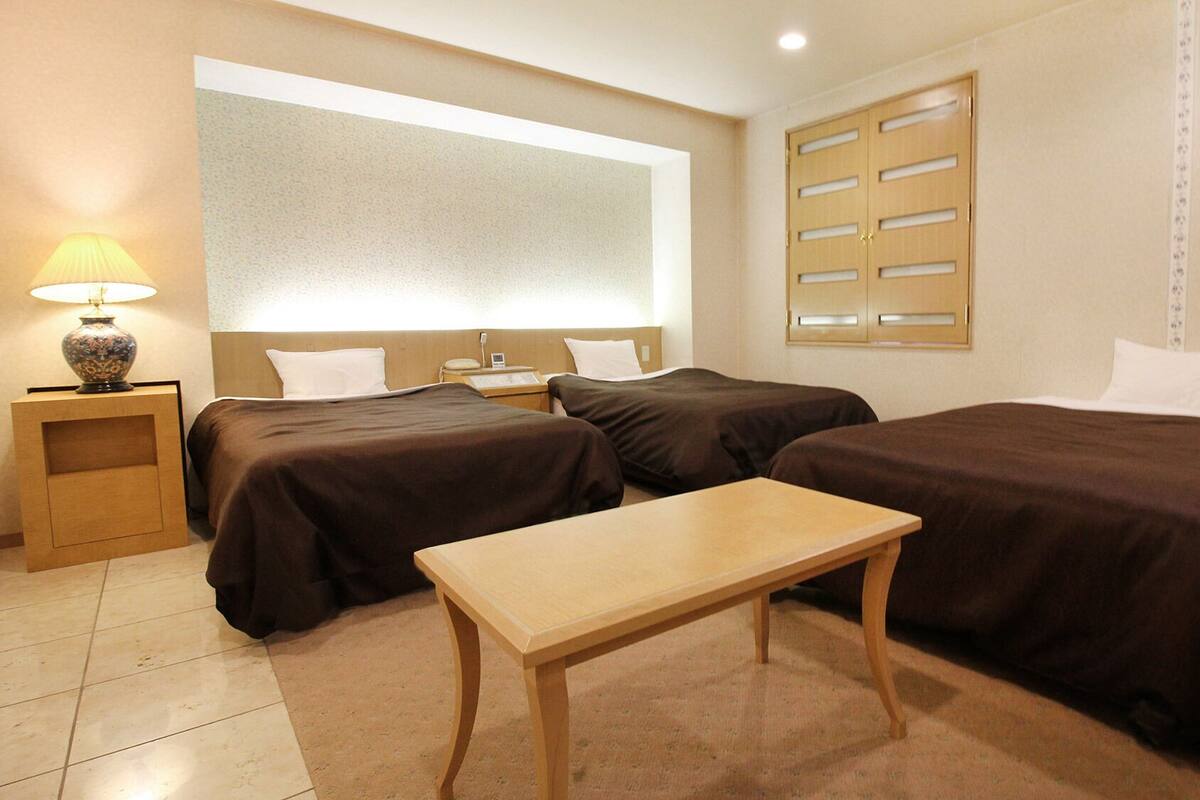 Une chambre d’hôtel avec deux lits, une petite table, une lampe et une fenêtre avec des stores.