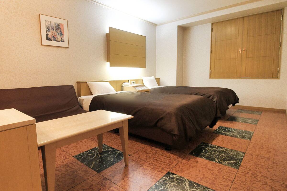Une chambre d’hôtel avec un lit, une table de chevet, une lampe, un tableau au mur et une fenêtre avec des stores.