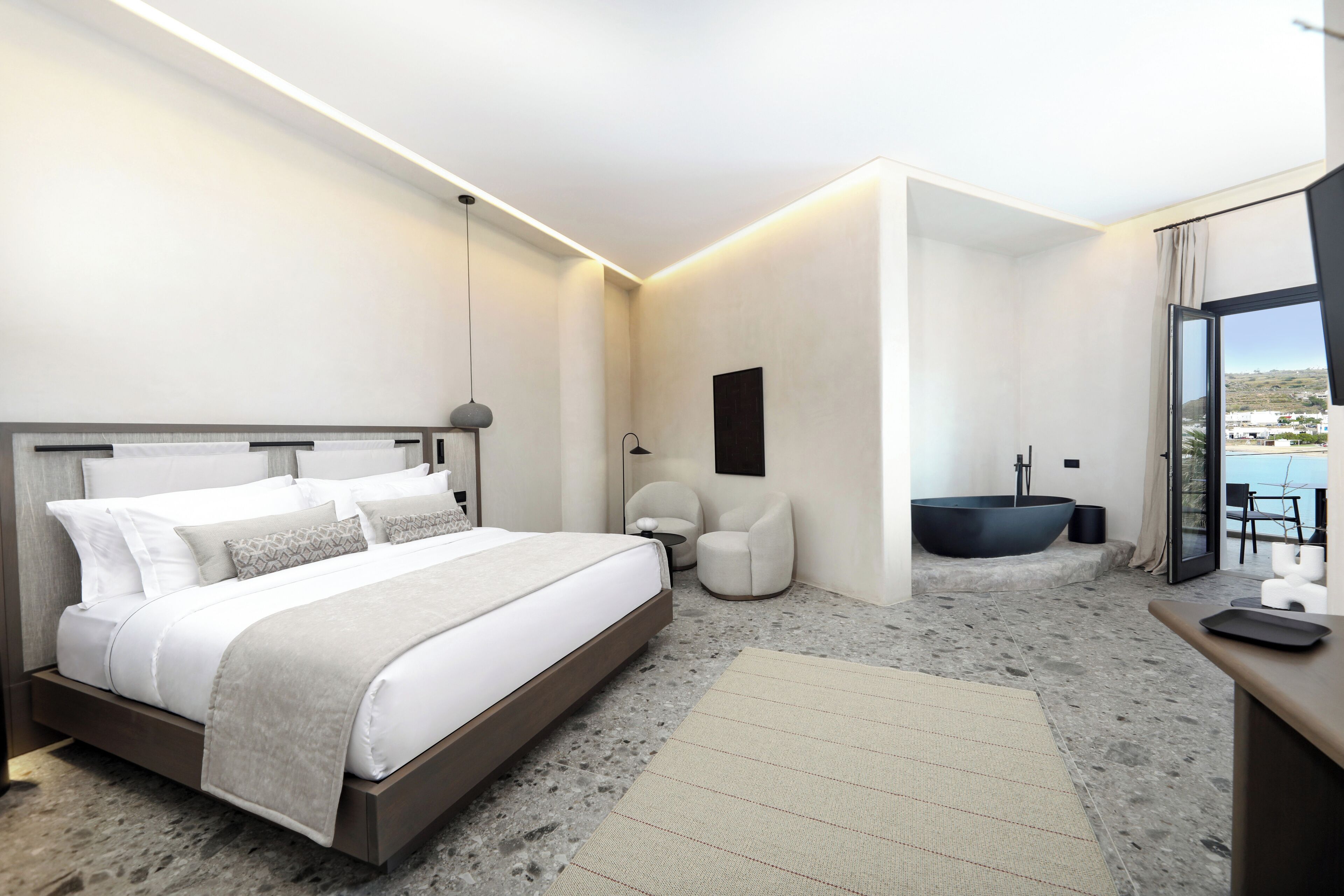 A modern hotel room with a large bed, a sitting area, a bathtub, and a view of the sea.