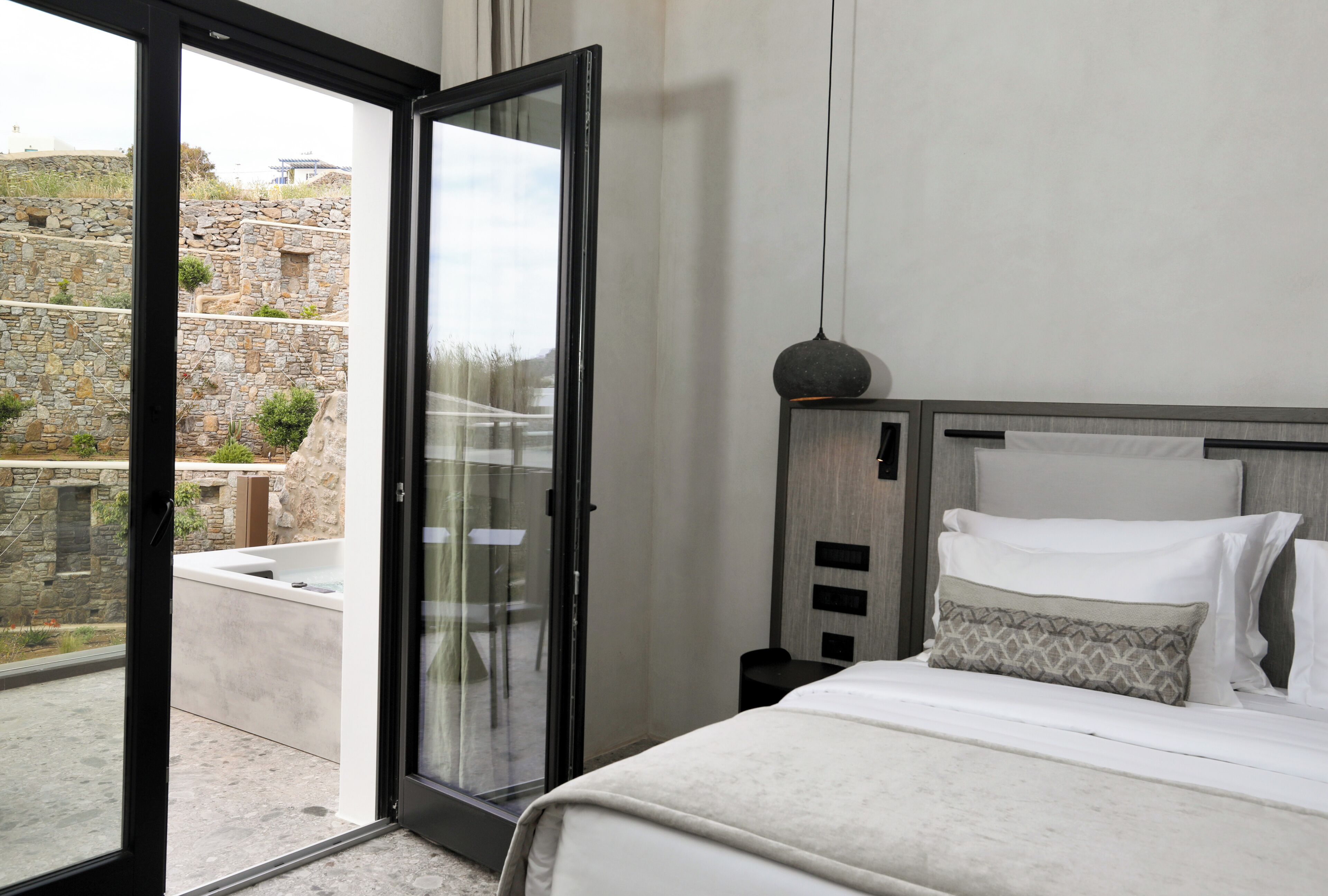 A bedroom with a bed, a nightstand, a bedside table, and a view through a glass door to an outdoor area with stone walls.