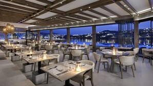 Un restaurante con mesas y sillas dispuestas para comer, amplios ventanales que ofrecen vistas de la ciudad de noche.