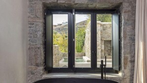 Un baño moderno con una bañera negra grande, una pared de piedra y vistas a una piscina y vegetación.