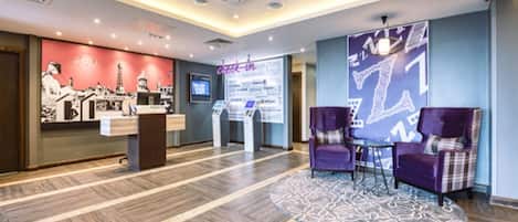 Un espace d’accueil d’hôtel comprenant un comptoir d’enregistrement, un écran numérique et un coin salon avec des fauteuils violets.