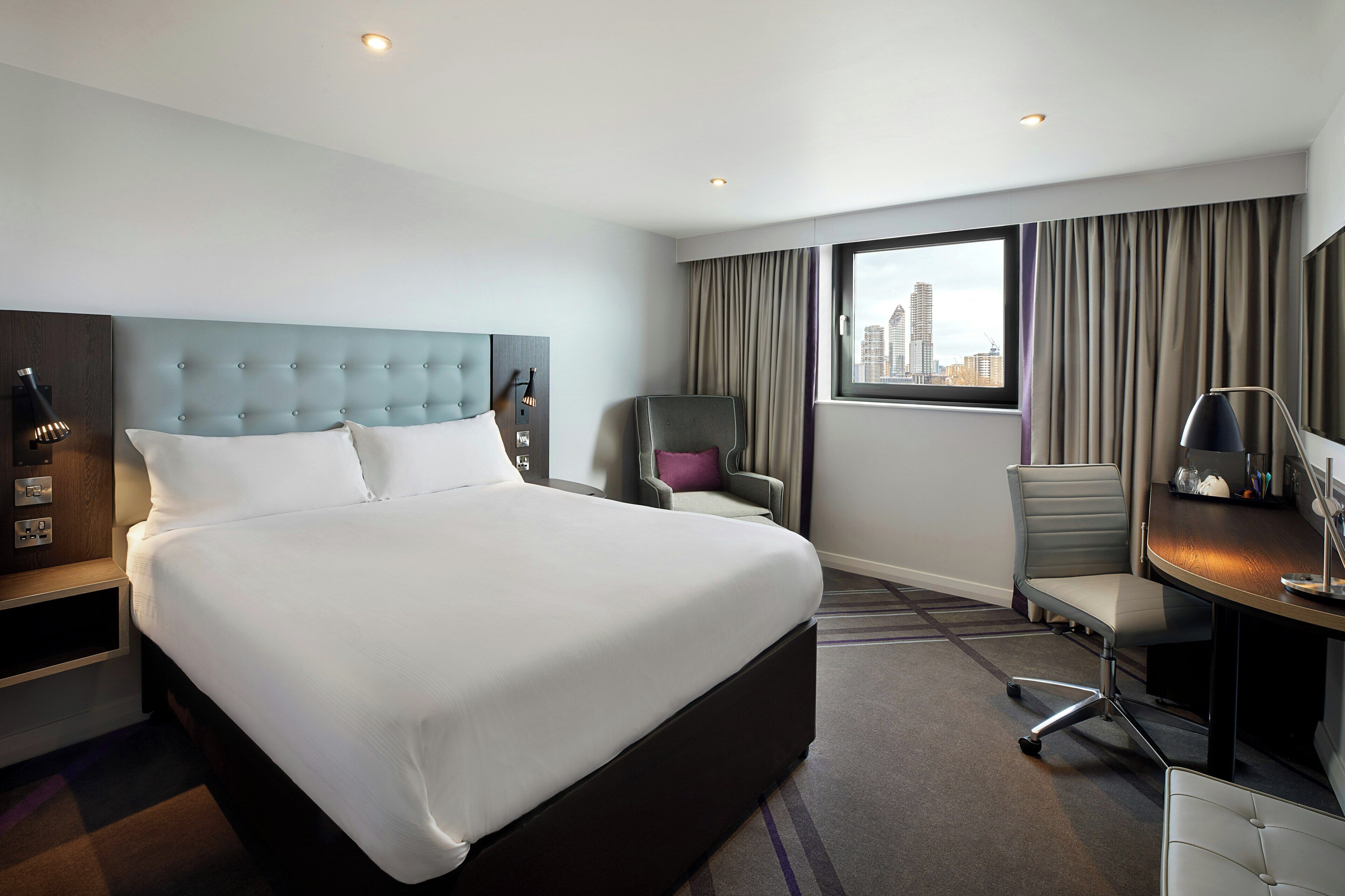 A hotel room with a large bed, a desk, a chair, and a view of a cityscape.
