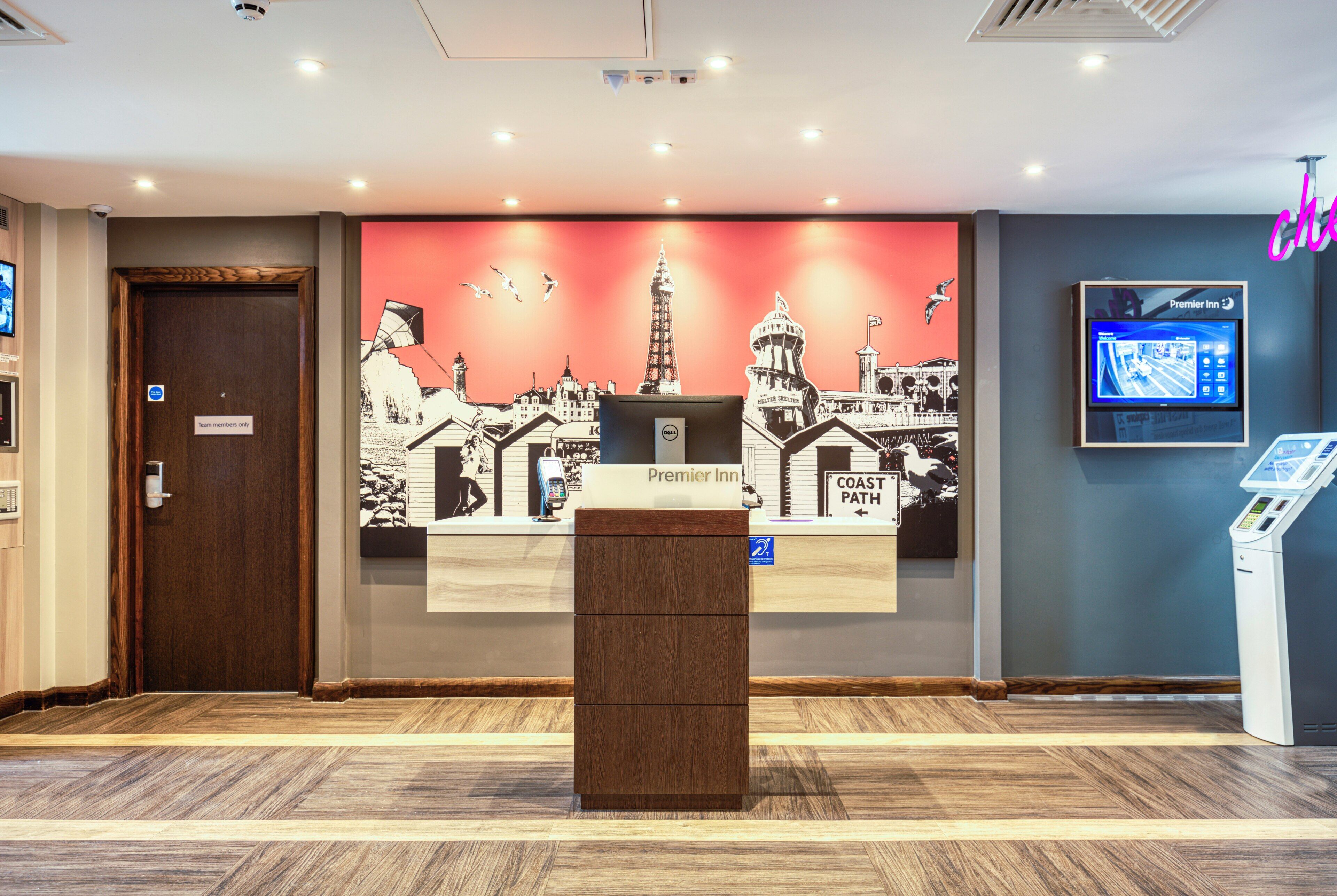 Uma área de recepção de hotel com uma tela digital, um balcão de atendimento e um mural com vista para a cidade.