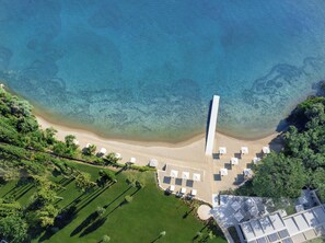 Vue aérienne d’un complexe hôtelier en bord de mer, avec une eau d’un bleu limpide, une plage de sable fin et des chaises longues et des parasols disposés avec soin.