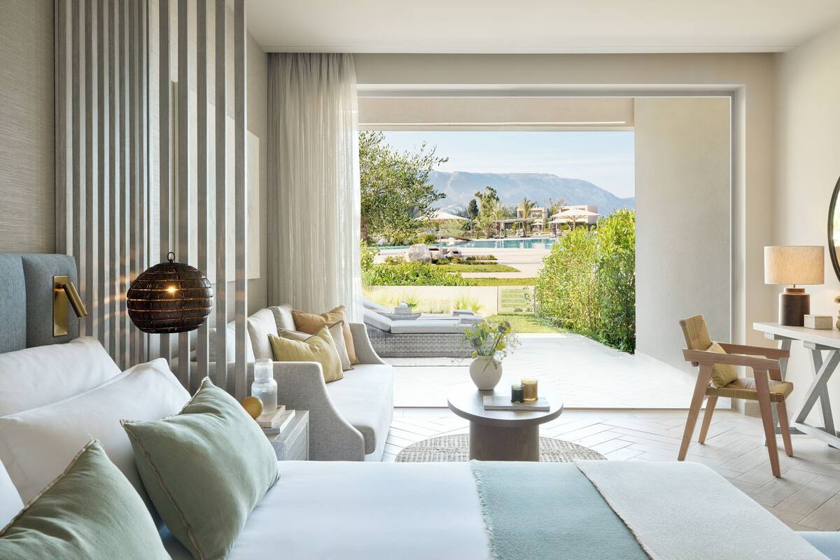 A modern hotel room with a large bed, a sofa, a dining table, and a view of a pool and mountains.