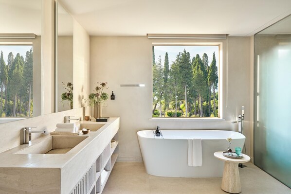 A modern bathroom with a freestanding bathtub, a large mirror, and a window with a forest view. - Ikos Odisia (Corfu)