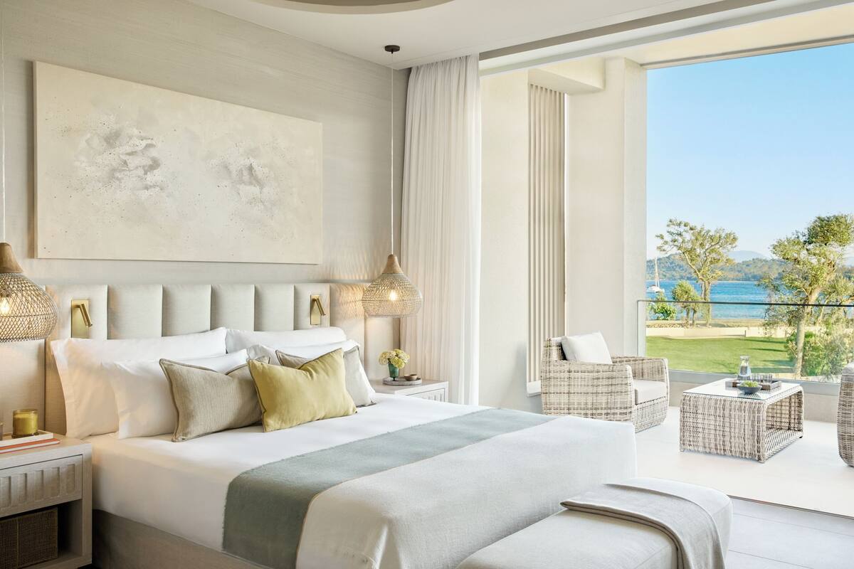 A modern hotel room with a large bed, a balcony with a view of the sea, and a large abstract painting on the wall.