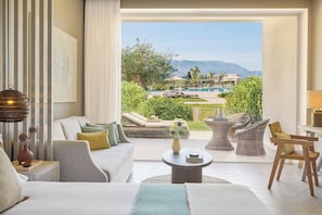 A hotel room with a large window, a sofa, a coffee table, and a view of a pool and mountains. - Ikos Odisia (Corfu)