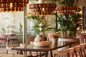 A dining area with a wooden table, wicker chairs, and a hanging light fixture with a circular design. - Ikos Odisia (Corfu)