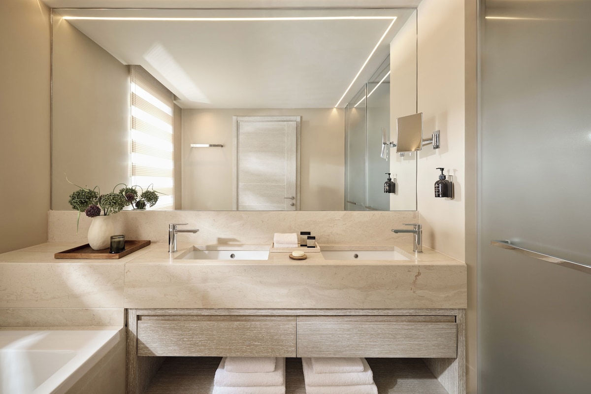 A modern bathroom with a large mirror, two sinks, a bathtub, and a wooden tray with a vase and candle.