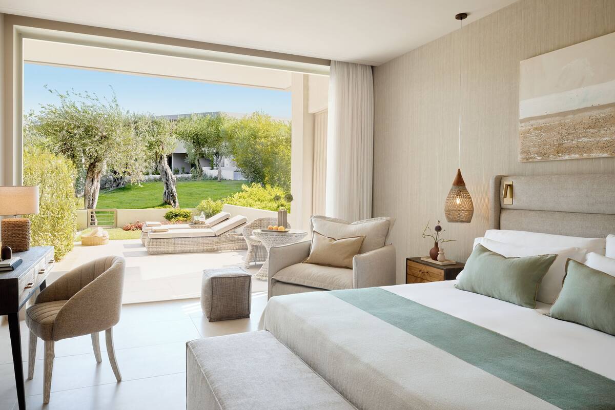A modern hotel room with a large bed, a seating area with a table, and a view of a garden.