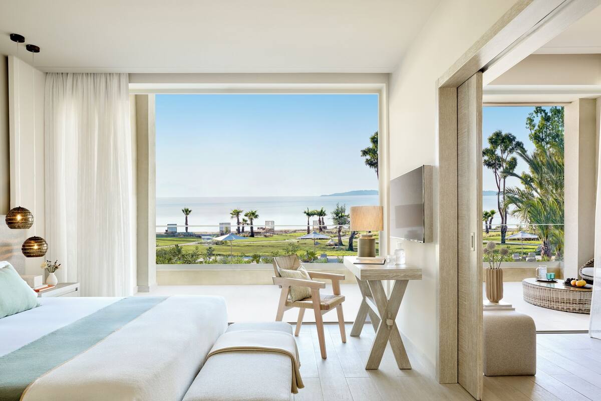 A hotel room with a large bed, a desk, a TV, and a balcony with a view of the sea.