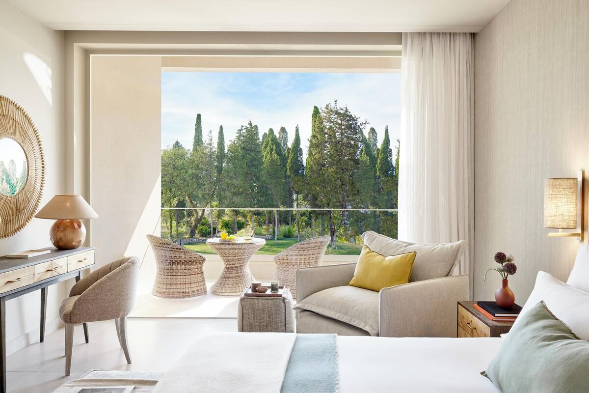 A modern hotel room with a balcony, a bed, a sofa, and a desk with a lamp.