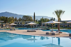 A resort pool area with lounge chairs, umbrellas, and a mountain backdrop. - Ikos Odisia (Corfu)