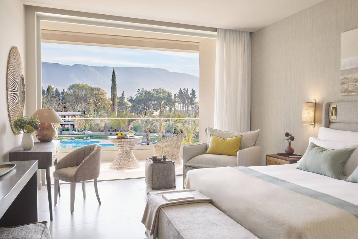 A hotel room with a bed, a desk, a chair, a small table, and a view of a pool and mountains.
