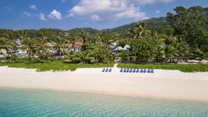 A beach with clear turquoise water, white sand, and blue lounge chairs. There are palm trees and houses in the background. - Laila, Seychelles, a Tribute Portfolio Resort (Mahé Island)