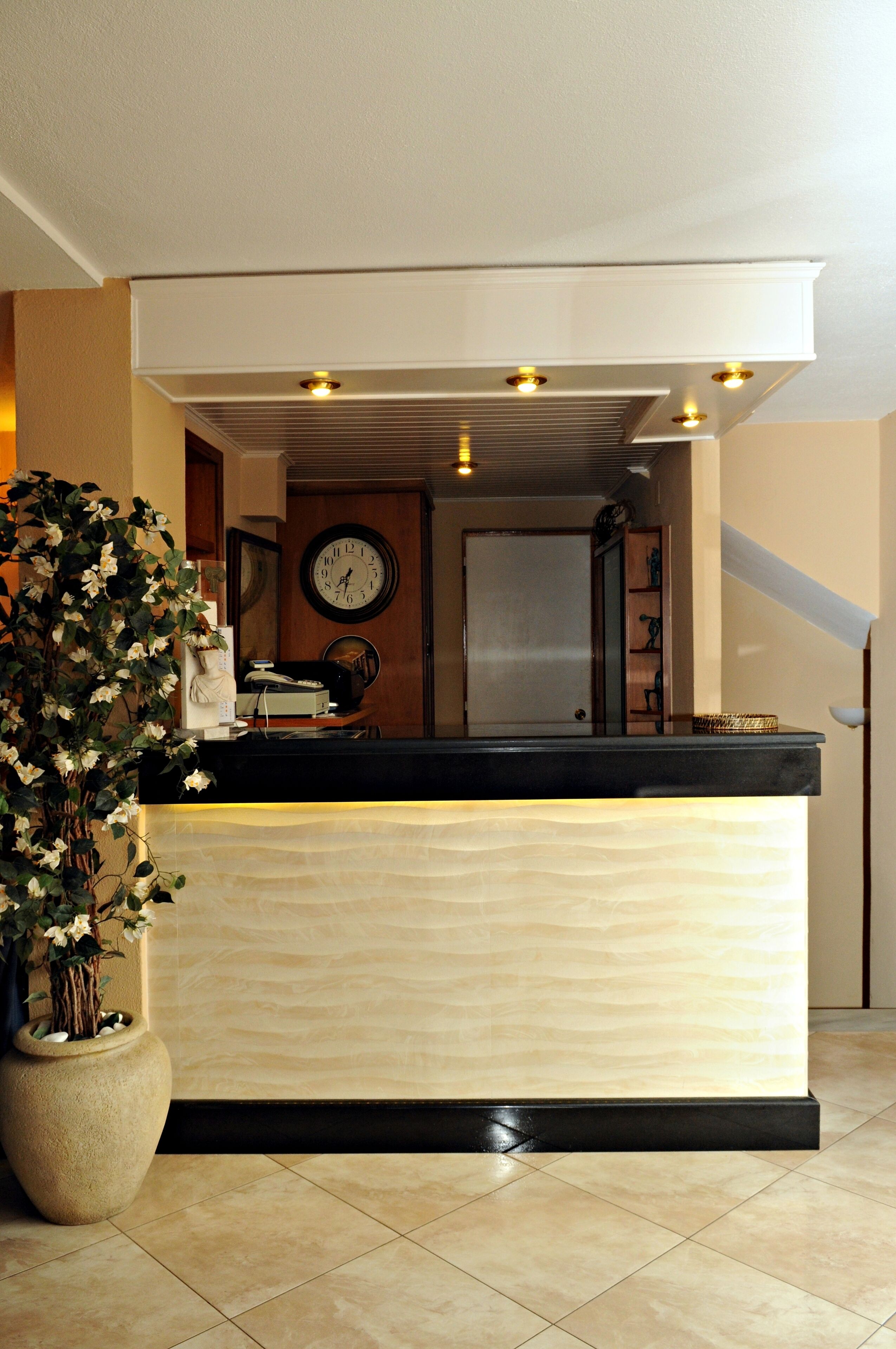 Ein Empfangsbereich mit einer Rezeption, einer großen Wanduhr, einem Blumentopf und einer Treppe im Hintergrund.