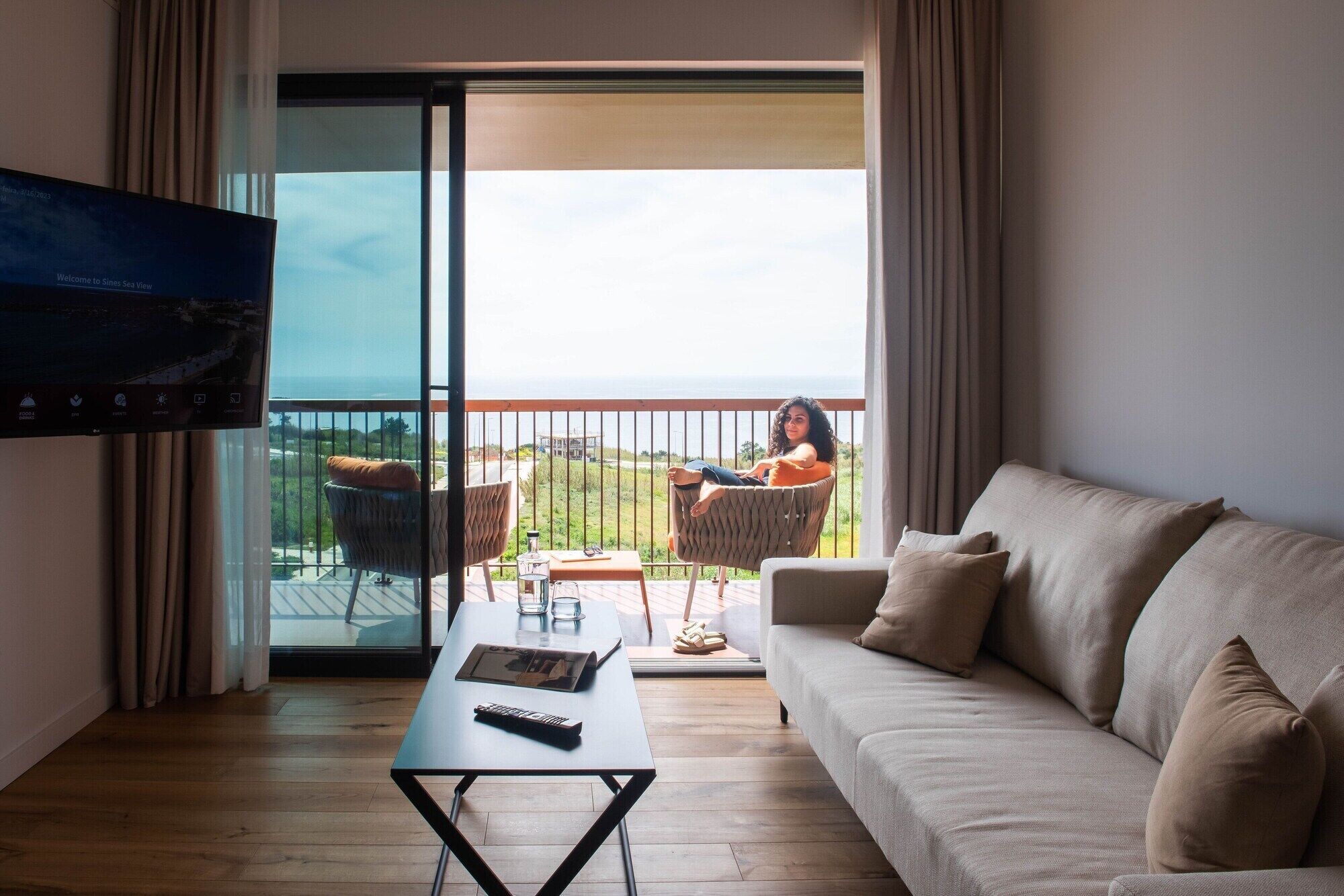 A room with a sofa, a table, a TV, and a balcony with a view of the sea.