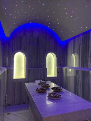 Una habitación con piso de mármol, una larga mesa con vajilla y tres ventanas arqueadas con interiores iluminados.