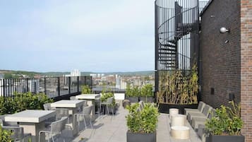 Um terraço ao ar livre com assentos e vasos de plantas, uma escadaria em espiral e vista para a paisagem urbana.
