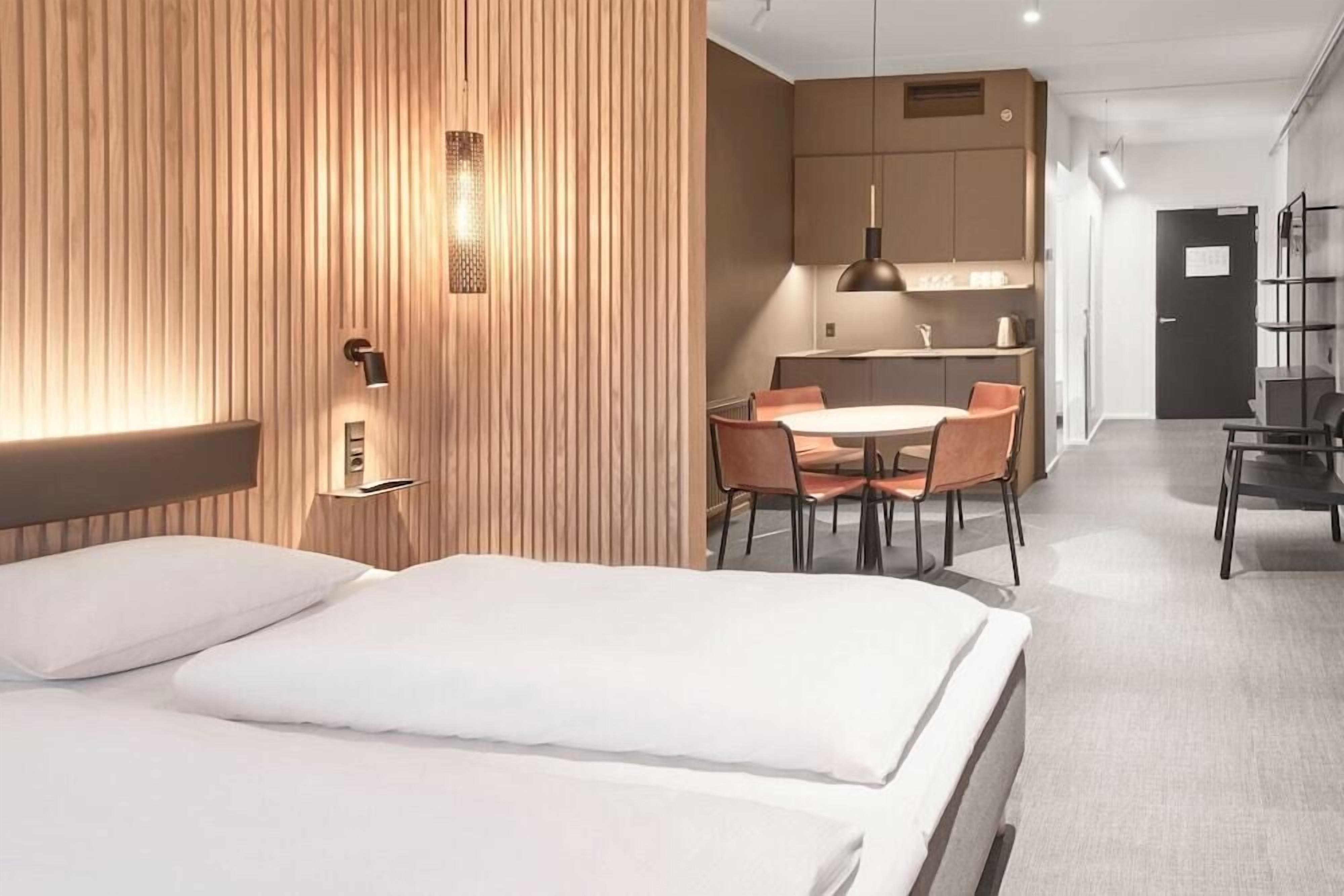 Quarto de hotel moderno com uma cama grande, parede decorativa em madeira, área de refeições com mesa e cadeiras, e uma cozinha compacta.