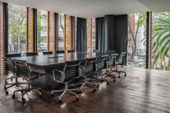 Une salle de conférence dotée d’une longue table et de chaises, de grandes fenêtres équipées de stores et offrant une vue sur les bâtiments et les arbres à l’extérieur.