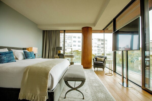 A modern hotel room with a large bed, a bench, a desk, and a view of the city through large windows. - Hotel Montevideo (Montevideo)