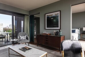 Una habitación de hotel moderna con un ventanal grande, zona de comedor, sofá, mesa de centro, cómoda y cama.