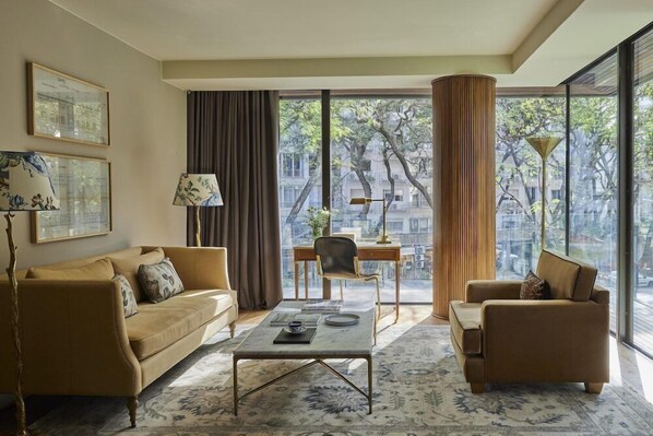 A modern living room with a sofa, armchair, coffee table, and a desk with a lamp. Large windows offer a view of buildings and trees. - Hotel Montevideo (Montevideo)