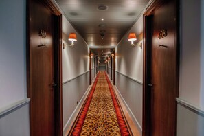 Un pasillo de hotel con puertas de madera, alfombra estampada y lámparas fijadas a la pared.